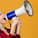 Funny excited young woman in checkered shirt shouting in megaphone over yellow background