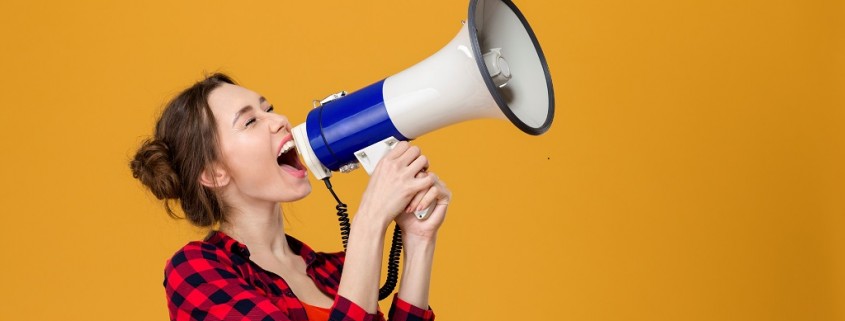 Funny excited young woman in checkered shirt shouting in megaphone over yellow background